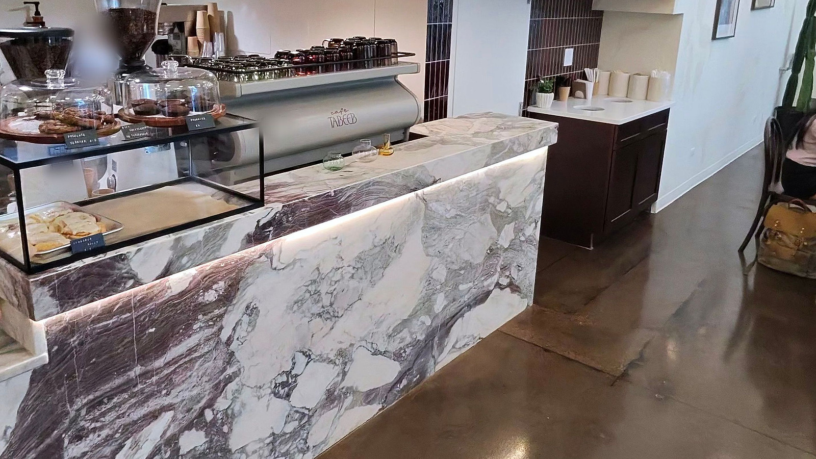 A finished cafe service counter clad in veined purple and white marble, lit from beneath the lip, with the espresso bar behind it