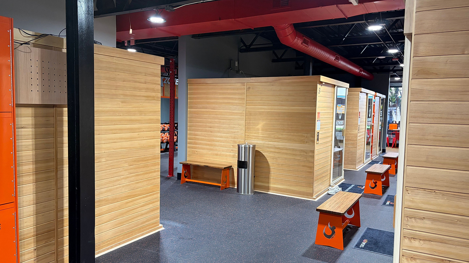 Cedar-clad infrared sauna enclosures on a fitness studio floor, with orange benches, exposed red ductwork and a black steel column