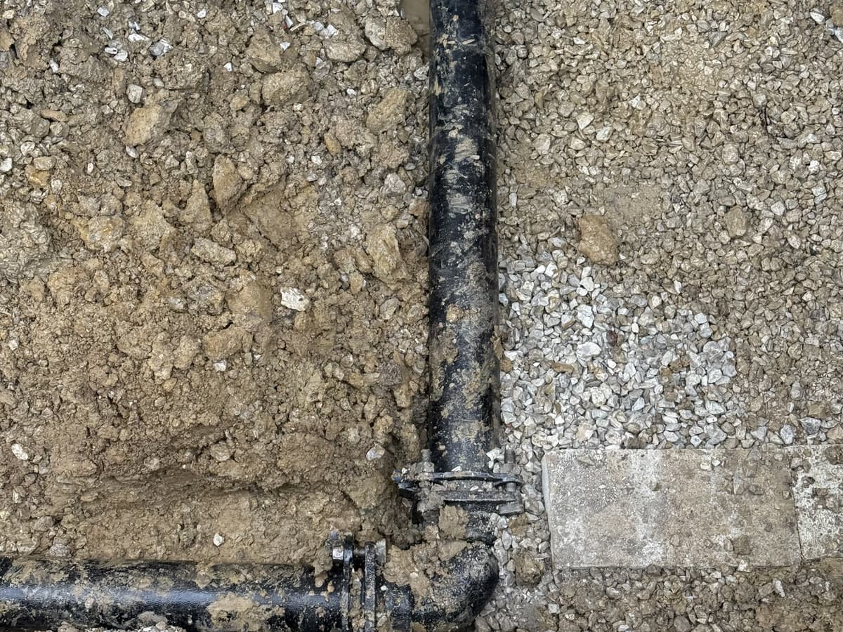 An open trench cut through a concrete slab with a new black pipe and fitting laid in it