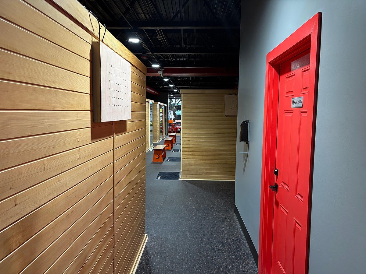 A corridor running between cedar-clad sauna enclosures, with a red-painted washroom door on the right and rubber flooring underfoot