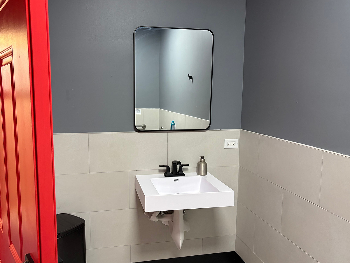 A washroom with large-format tile to mid-height under a gray painted wall, a wall-hung basin with black fittings and a mirror