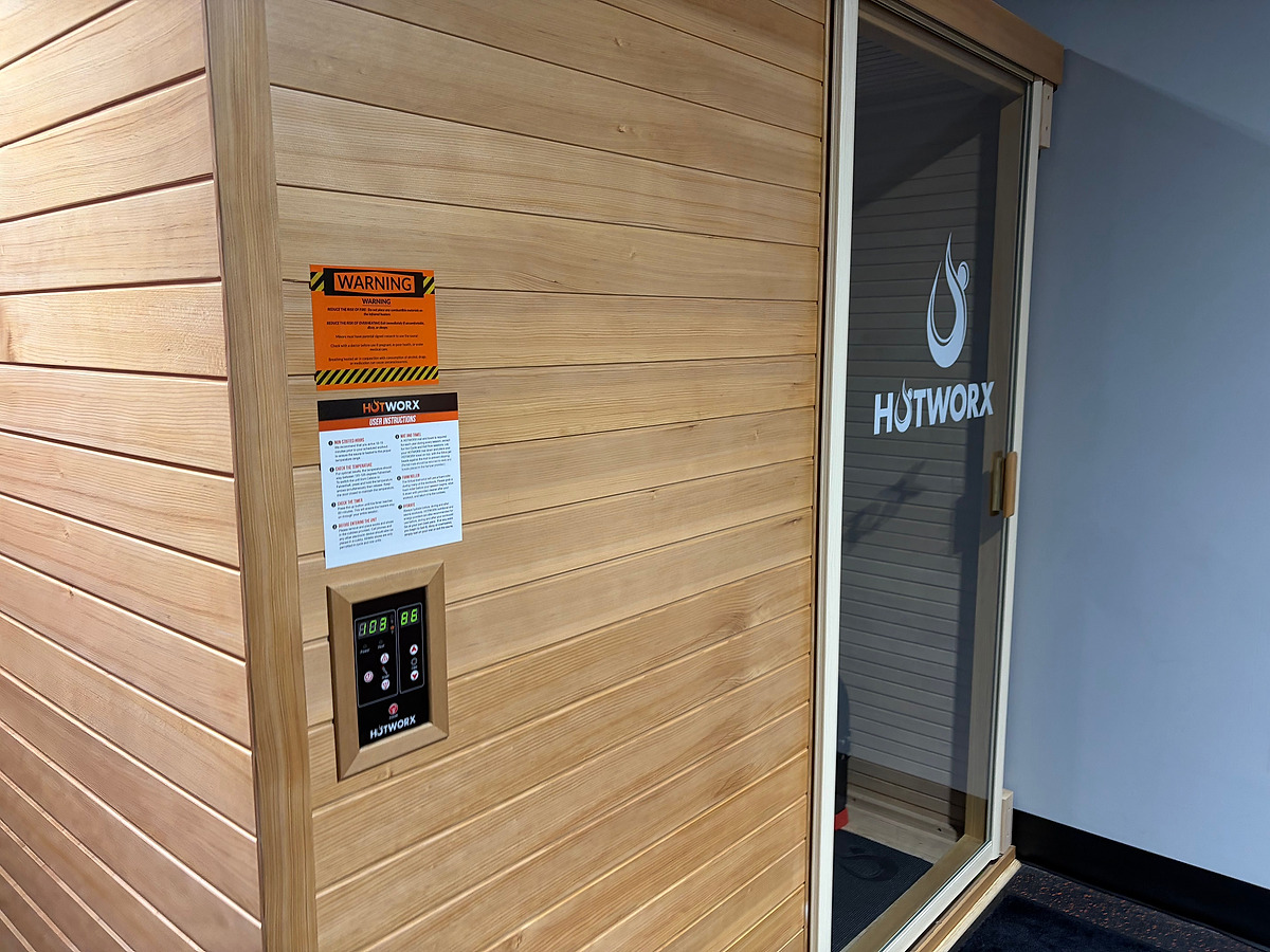 A cedar-clad sauna with the HOTWORX name on its glass door, a wall-mounted control panel and posted instructions beside it