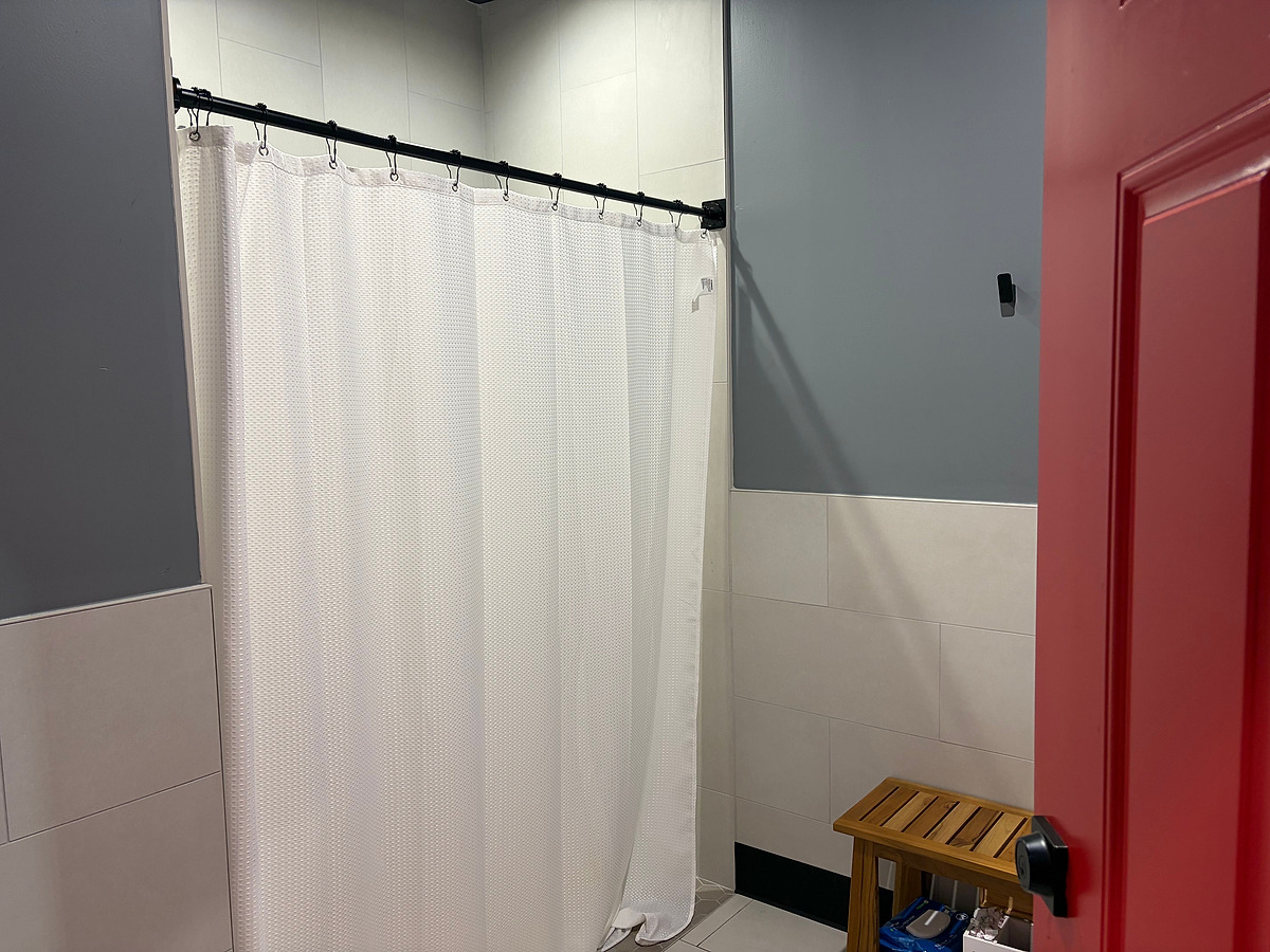 A shower stall behind a white curtain on a black rail, tiled to full height, with a gray painted wall and a red door leaf