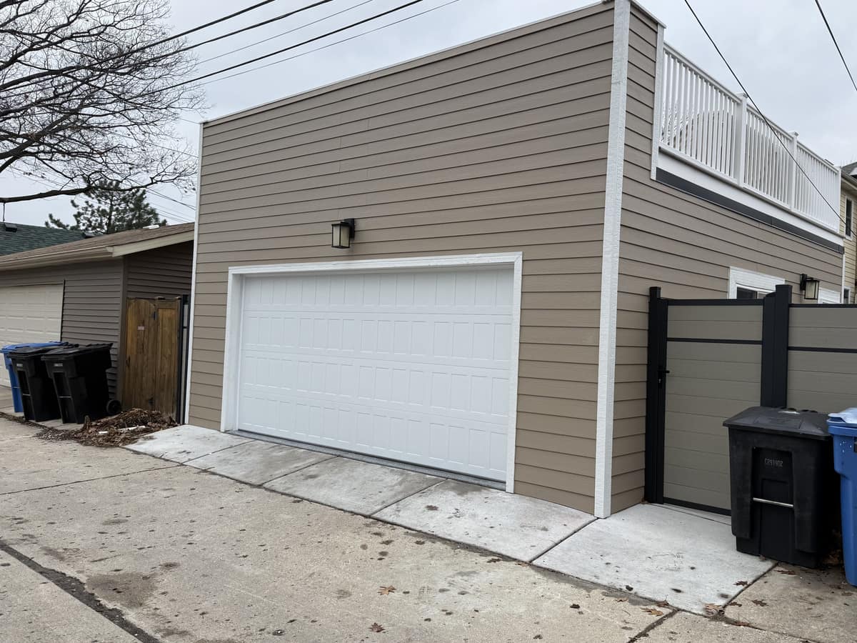 A detached garage with tan lap siding and a white double garage door, a railed roof deck above, seen from the alley