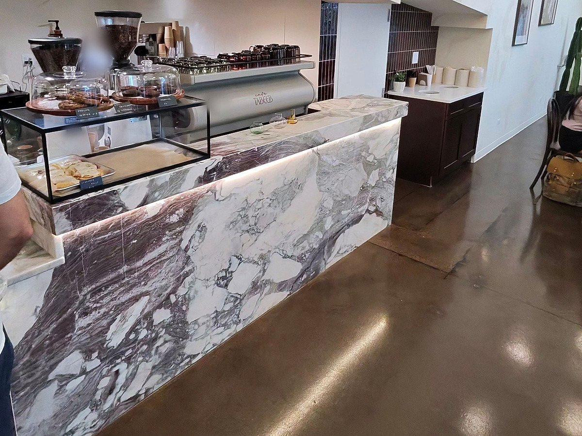 A cafe service counter clad in veined purple and white marble, lit from beneath the lip, with a pastry case and espresso bar behind it