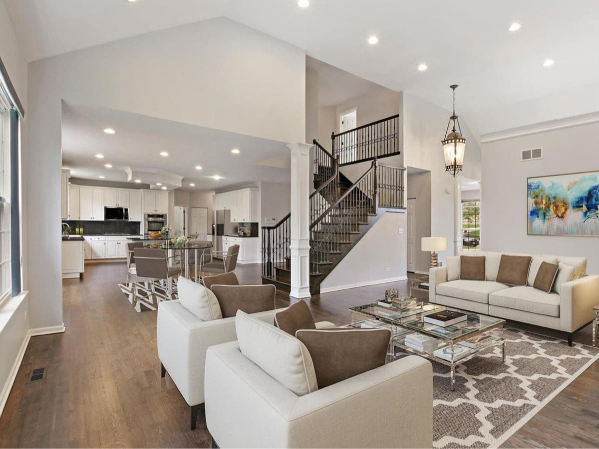 A furnished open-plan living room under a vaulted ceiling, with wide-plank hardwood and an open stair with dark treads