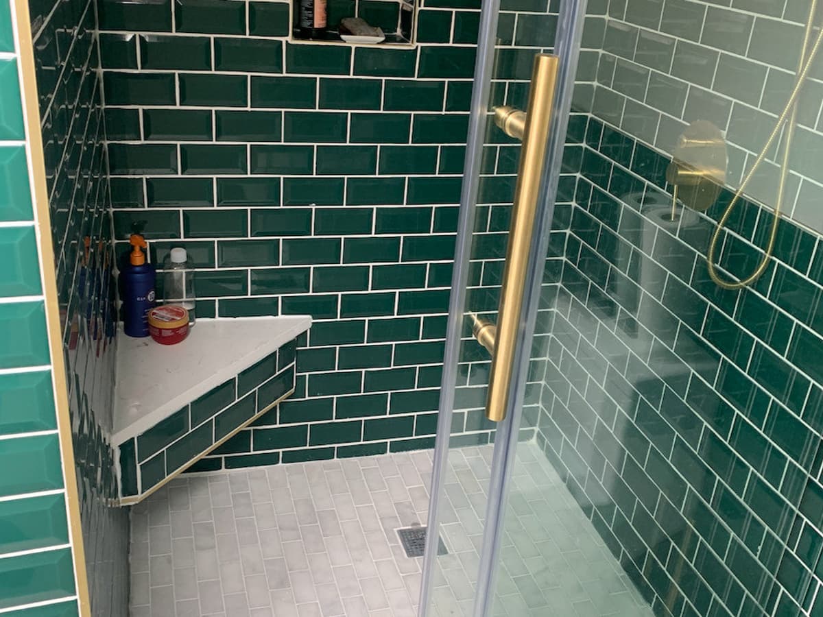 A walk-in shower lined in dark green beveled subway tile, with brass fittings and a built-in corner bench
