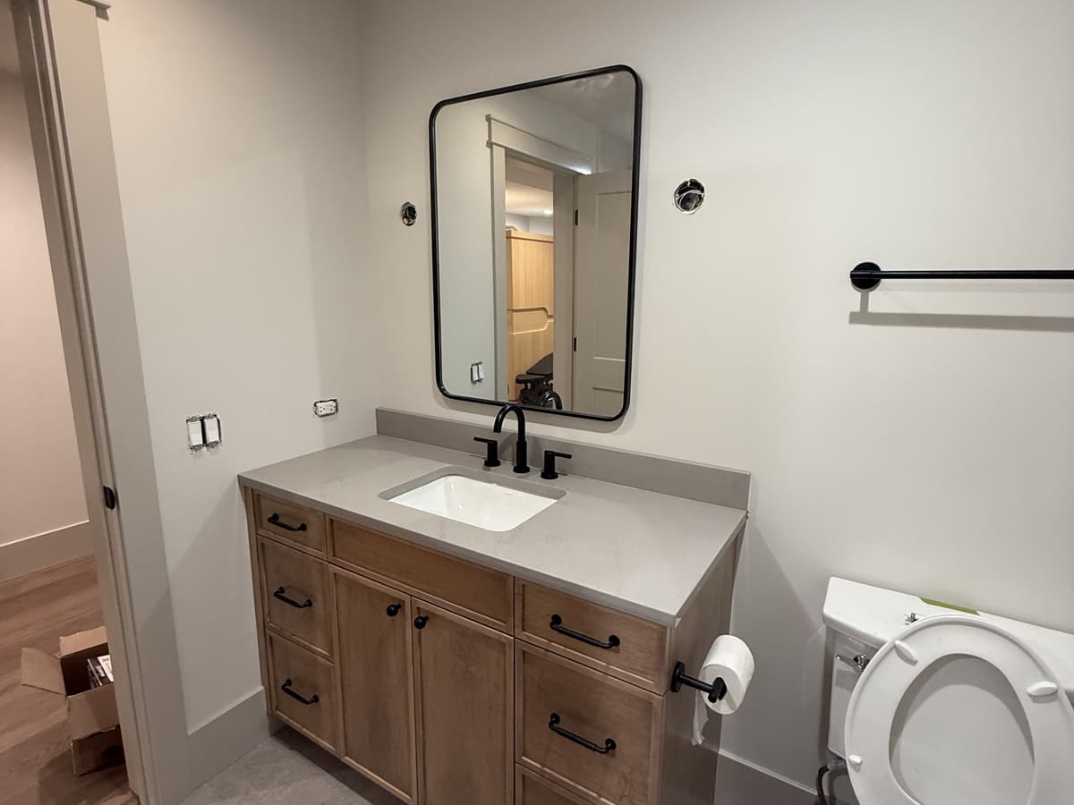 A bathroom vanity in warm wood with a white top, a black framed mirror and black fittings