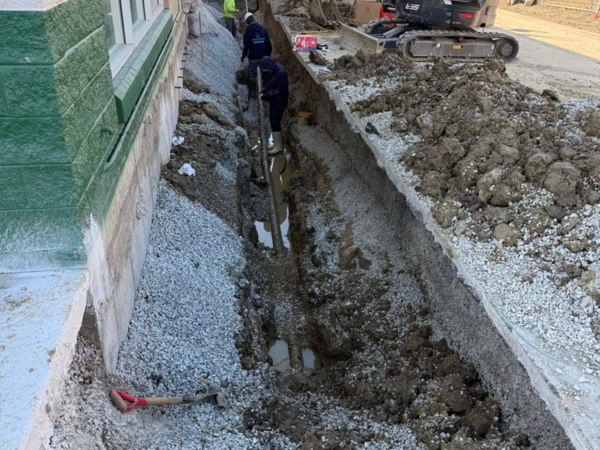 A narrow trench excavated along a building foundation, with a worker in the trench and an excavator behind