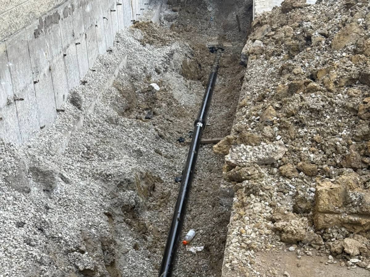 Black pipe bedded in the base of a trench alongside a foundation wall, with a coupling and a branch fitting
