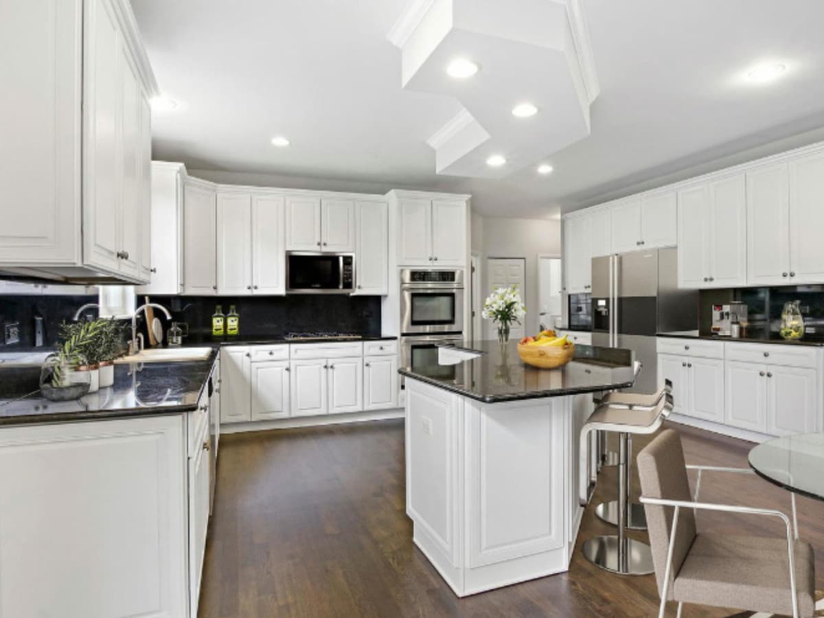 A white kitchen with a stone-topped island and stools, under a vaulted ceiling with recessed lighting